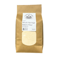 A 1kg bag Organic Whole Heritage Bakers Flour, shown in a brown kraft paper bag with a white label and a clear window displaying the pale flour inside.